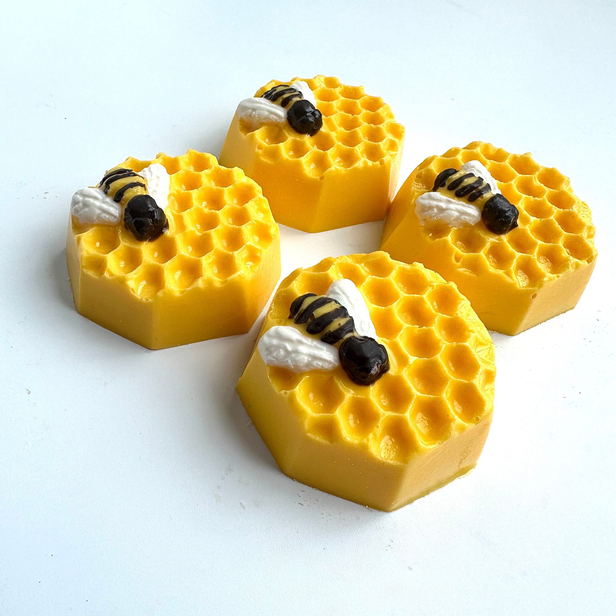 Four hexagonal, yellow beeswax soaps with honeycomb patterns and bee decorations are arranged on a white surface.