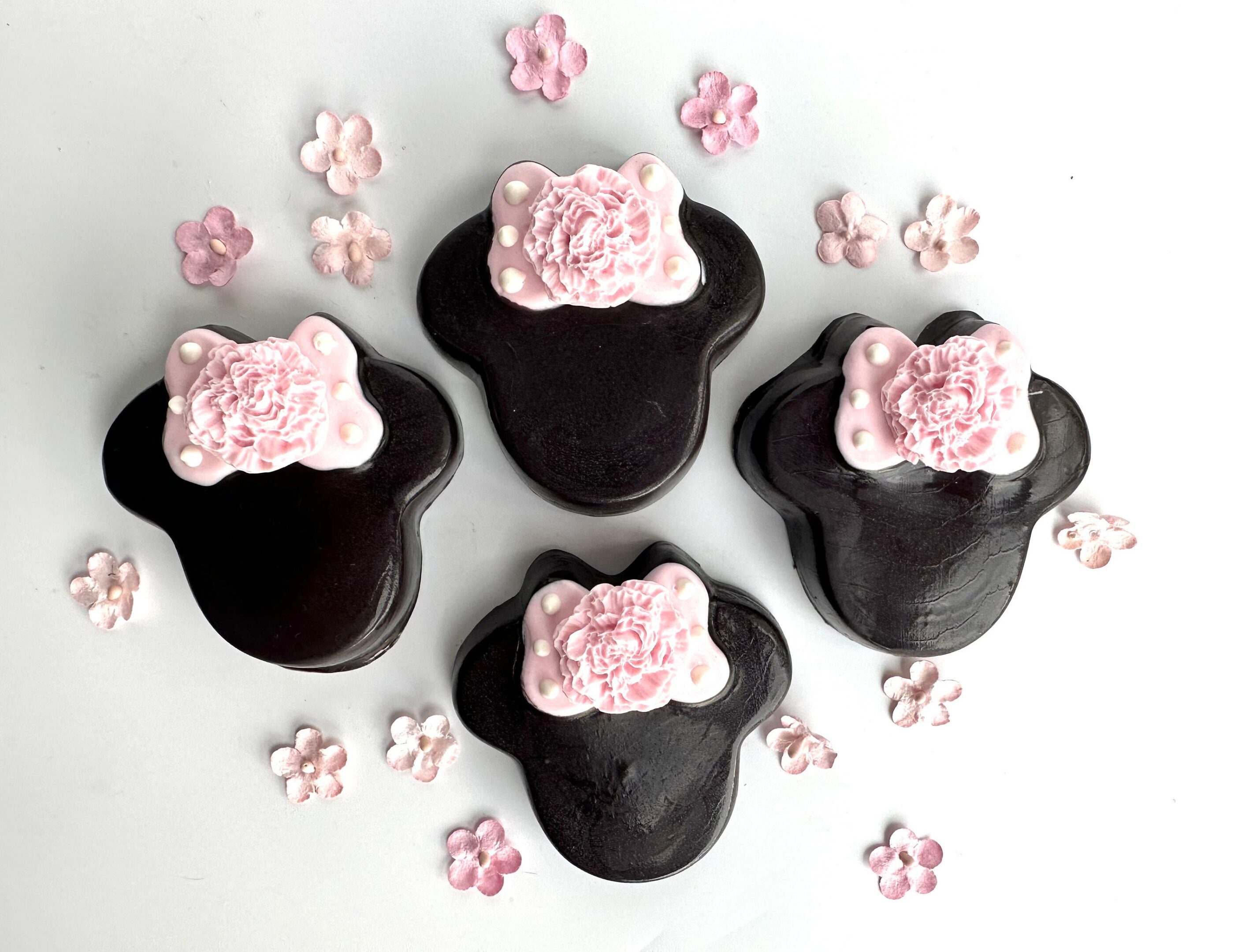 Four bear-shaped Minnie Chocolate Covered Oreos with pink floral icing details, surrounded by small pink flower decorations on a white background.