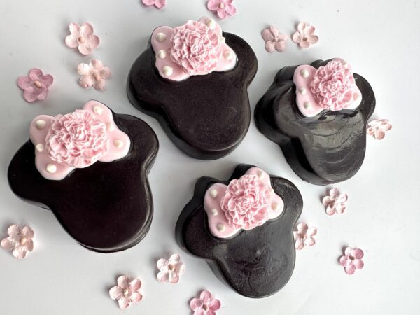 Four Minnie Chocolate Covered Oreos, shaped like black bears with pink floral decorations, surrounded by small pink flowers on a white background.