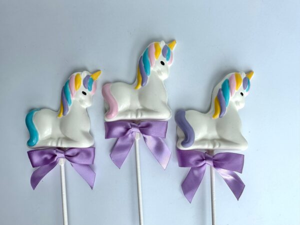 A set of three unicorn-shaped white chocolate lollipops features colorful manes and golden horns, adorned with purple ribbons, displayed against a plain white background.