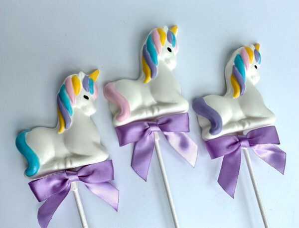 A trio of unicorn white chocolate lollipops with colorful manes and tails, each tied with a purple ribbon, for parties like baby showers or birthdays.