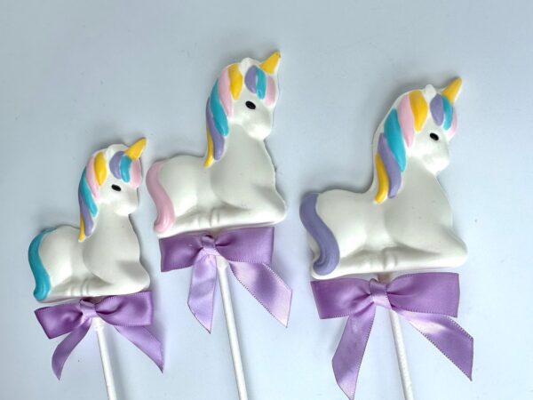 Three unicorn-shaped white chocolate lollipops, part of a 24-piece set for baby showers and parties, feature pastel manes and horns with purple bows against a plain white background.