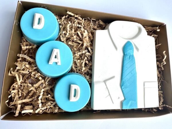 Gift box with a chocolate-covered Oreo, three blue circular chocolates spelling "DAD," and a white chocolate shirt accessorized with a blue tie, all nestled in shredded paper.