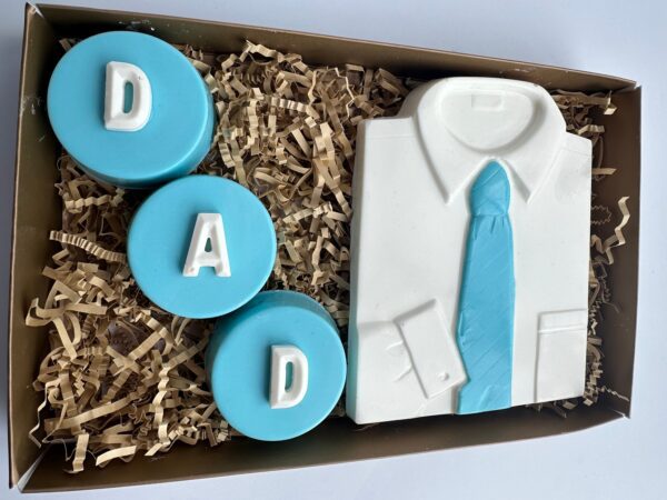 Gift box containing a white chocolate shirt with a blue tie and three round blue chocolates spelling "DAD," arranged on shredded paper. Product: DAD Chocolate Covered Oreo and White Chocolate Shirt Gift Box.