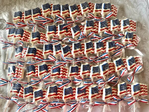 Rows of American flag-shaped white chocolate lollipops, perfect for a Memorial Day or 4th of July picnic, each tied with a red, white, and blue ribbon on a patterned background.