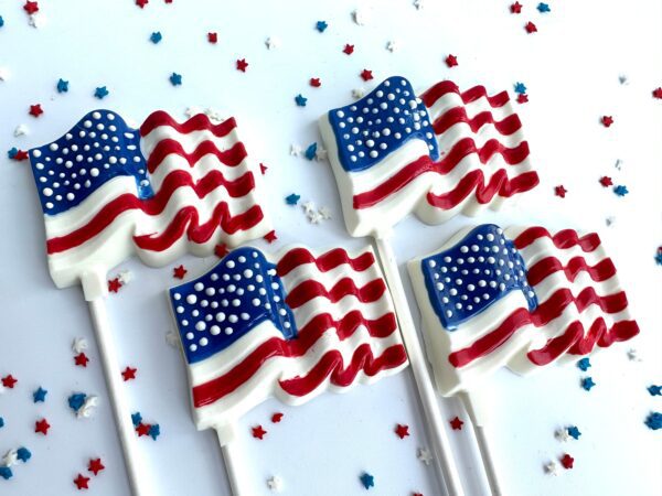 Five white chocolate lollipops with an American flag design are displayed on a white surface, surrounded by scattered red, white, and blue star-shaped decorations.