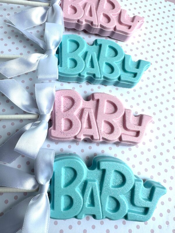 Four "BABY" chocolate lollipops, adorned with white ribbons on sticks, are arranged neatly on a dotted surface for baby showers and party favors.