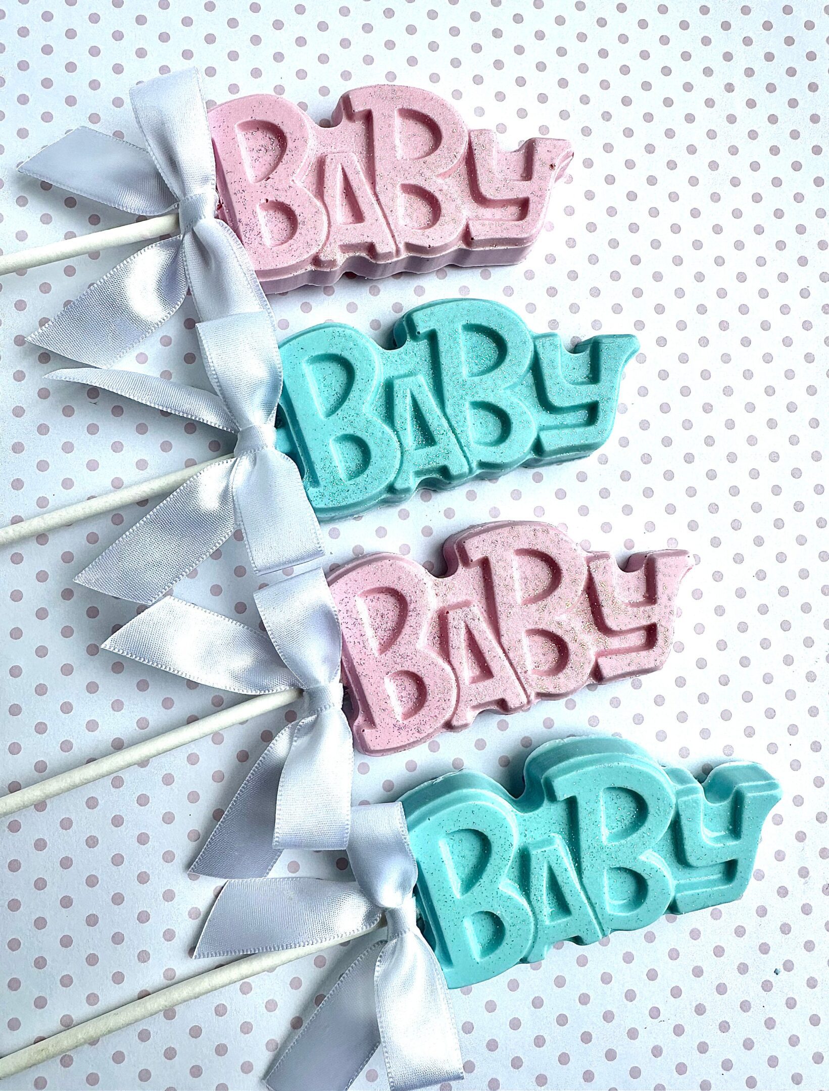 24-BABY Chocolate Lollipops for Baby Showers, featuring pink and blue designs with white bows on sticks, set against a polka dot background.