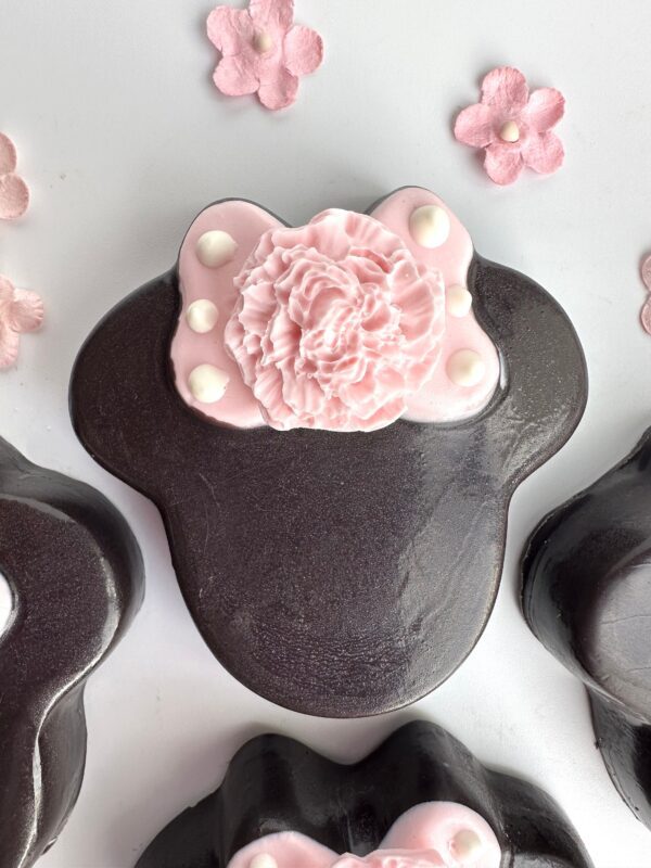 A set of 12 Minnie-themed chocolate-covered Oreos featuring black Mickey Mouse shapes with pink carnations, white dots on top, and adorned with small pink flower decorations—perfect for party favors or celebrations like birthdays and baby showers.