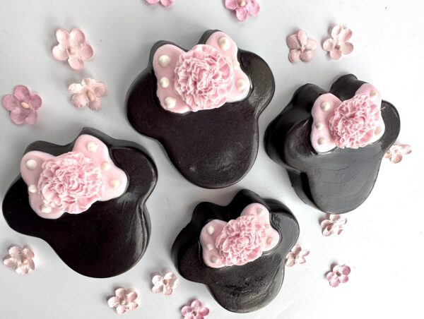 Four chocolates shaped like black bear paws, featuring pink floral decorations and surrounded by small pink flowers on a white background. Ideal for party favors or celebrations like birthdays and baby showers; part of the 12-Minnie Chocolate Covered Oreos collection.