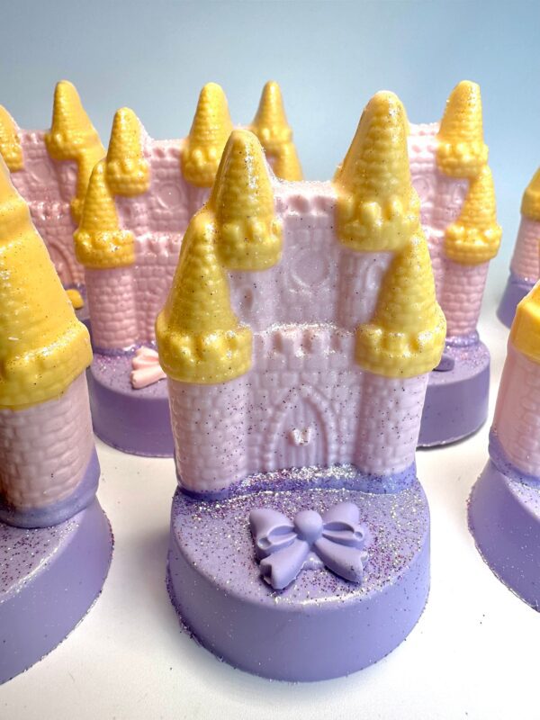 Twelve pink and yellow castle-shaped decorations sit on purple, glittery bases with bow details; these are the 12-Fairytale Castles on Chocolate Covered Oreos ideal for a Princess, Rainbow, or Unicorn Party.