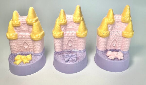 A set of three pink soap molds shaped like fairytale castles with yellow turrets and decorative bows, resting on a purple base.