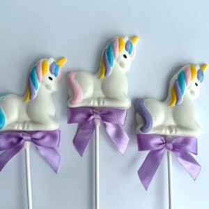 Three unicorn-shaped white chocolate lollipops, adorned with multicolored manes, tails, and purple ribbons from the "24-Unicorn White Chocolate Lollipops For Baby Shower/Birthday Party/Party Favors" collection, are placed on a white background.