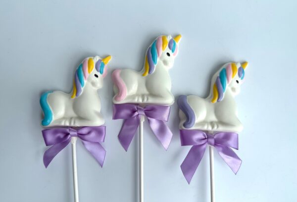 Three unicorn-shaped white chocolate lollipops, adorned with multicolored manes, tails, and purple ribbons from the "24-Unicorn White Chocolate Lollipops For Baby Shower/Birthday Party/Party Favors" collection, are placed on a white background.