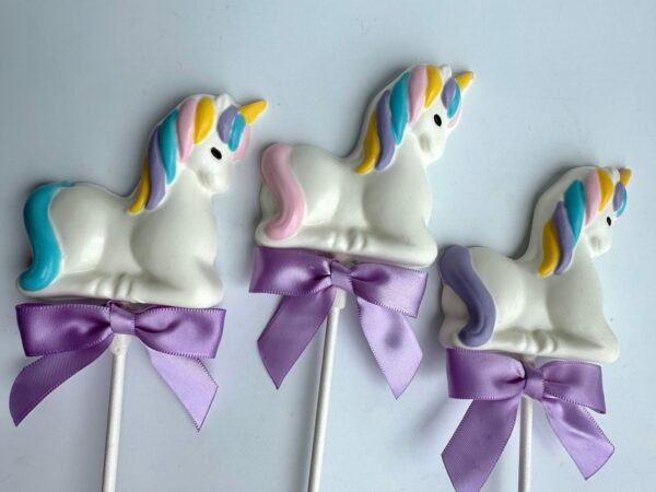 Three white unicorn-shaped chocolate lollipops from the 24-Unicorn White Chocolate Lollipops set, featuring colorful manes and purple bows, are arranged in a row on a white background.
