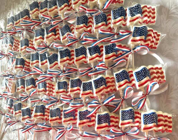 Rows of white chocolate lollipops, each decorated as an American flag and tied with red, white, and blue ribbons, arranged on a patterned surface.