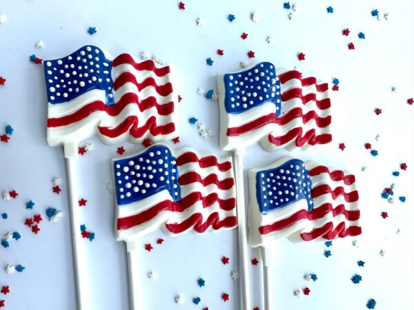 Five out of a set of 12 white chocolate American flag-shaped lollipops with red, white, and blue details are on a white surface surrounded by small star-shaped confetti.