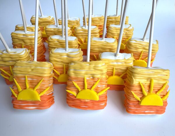 Sunset Ombré Rice Krispie Treats are rectangular cakes on sticks, decorated with yellow and orange icing in a sunset theme with sun designs, perfect for birthdays or baby showers.