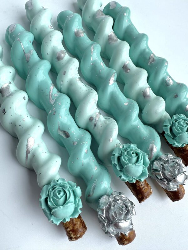 These are 12 caramel pretzel sticks, coated in teal and white chocolate with silver accents, and decorated with flower shapes on one end, perfect for weddings, showers, birthdays, or party favors.