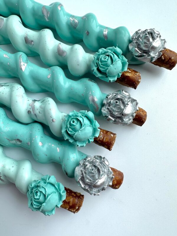 Five Tiffany Blue and silver chocolate-covered caramel pretzels with floral decorations on a white background.
