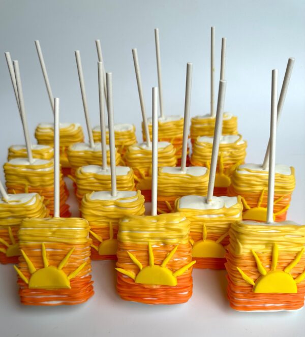 Several Sunset Ombré chocolate-covered rice krispie treats are decorated with yellow and orange swirls, featuring a flat fondant sun design on the front, and have white sticks.