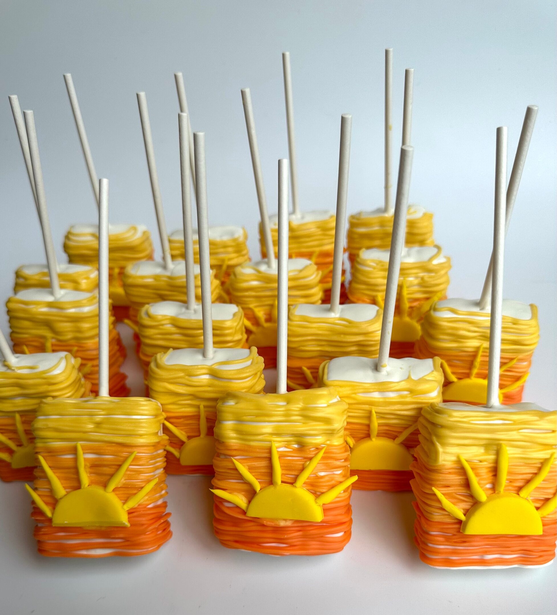 Several Sunset Ombré chocolate-covered rice krispie treats are decorated with yellow and orange swirls, featuring a flat fondant sun design on the front, and have white sticks.