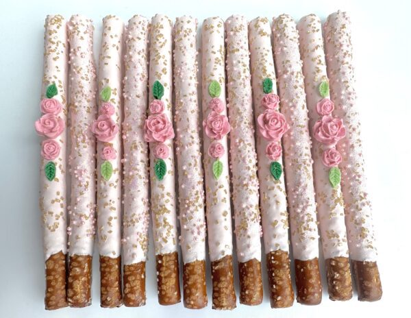 Twelve pale pink chocolate-covered pretzels, ideal for birthdays, weddings, or showers, adorned with pink roses, green leaves, and gold sprinkles.