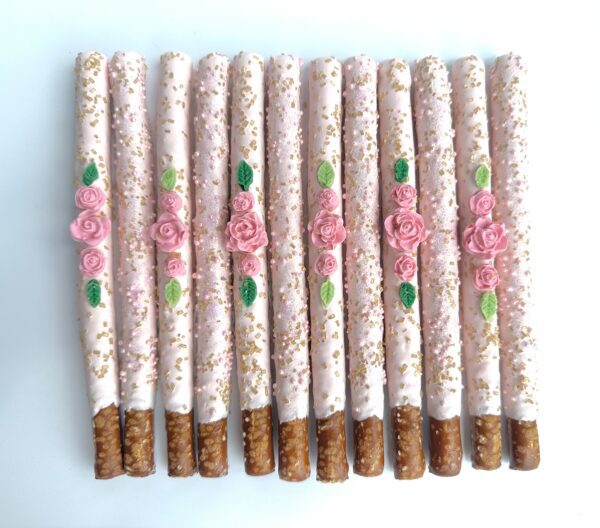 Twelve pale pink chocolate-covered pretzels, decorated with roses and leaves, are neatly arranged on a white surface. Perfect for birthday parties, wedding favors, or shower favors.