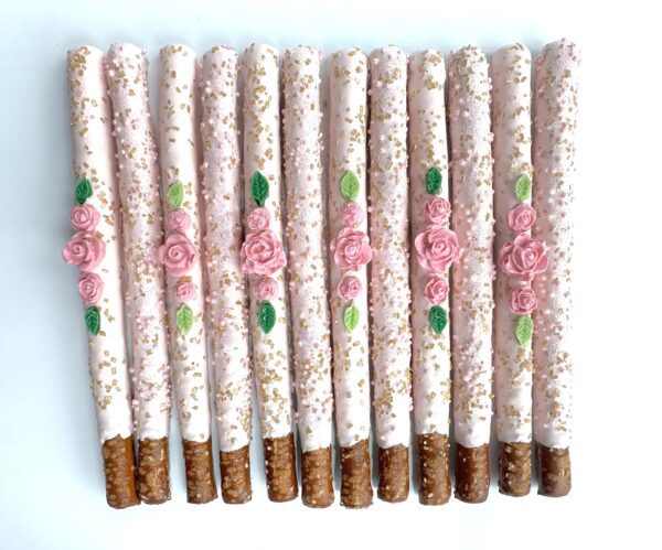 Twelve pale pink chocolate-covered pretzel sticks, perfect for birthday parties, weddings, or shower favors. Decorated with pink roses and gold sprinkles, these pretzels add a touch of elegance to any occasion.
