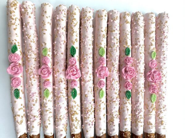 12 pale pink chocolate-covered pretzels, decorated with roses, leaves, and gold sprinkles, lined up on a white surface for birthday parties or wedding/shower favors.