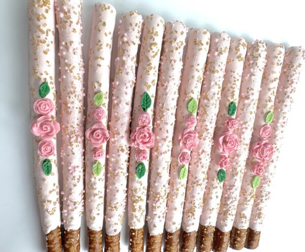 A row of 12 pale pink chocolate-covered pretzels with gold sprinkles, adorned with small pink roses and green leaves, perfect for birthdays, weddings, or showers.