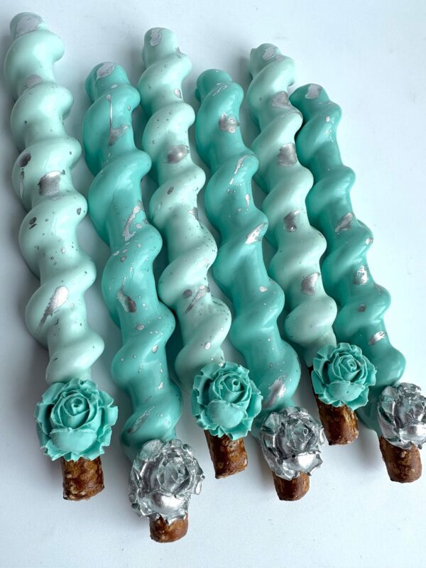 Six twisted pretzel sticks are coated in white, light blue, and teal chocolate with metallic accents and floral decorations on a white background.