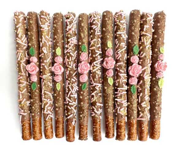 12-Dainty Milk Chocolate Covered Pretzels decorated with pink sprinkles, gold sparkles, and pink rose icing are arranged in a row. Perfect for Birthday Party/Wedding Favor/Shower Favors.