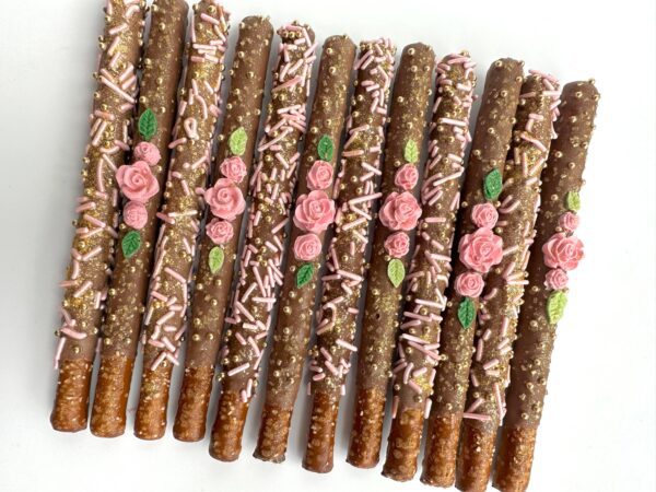 Nine of the "12-Dainty Milk Chocolate Covered Pretzels With Flowers For Birthday Party/Wedding Favor/Shower Favors" are decorated with pink sprinkles and rose embellishments, arranged in a row on a white surface.