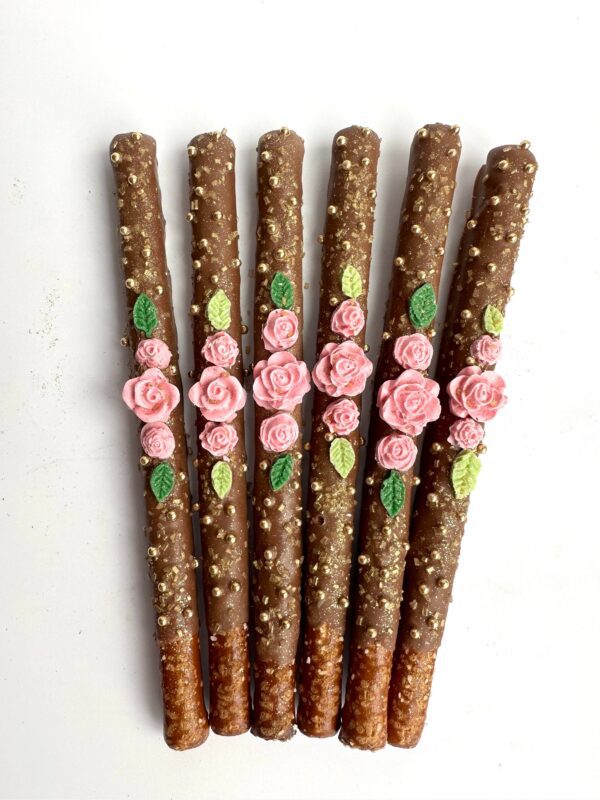 Twelve milk chocolate-covered pretzel sticks, adorned with floral icing designs in pink and green, are fanned out on a white background.