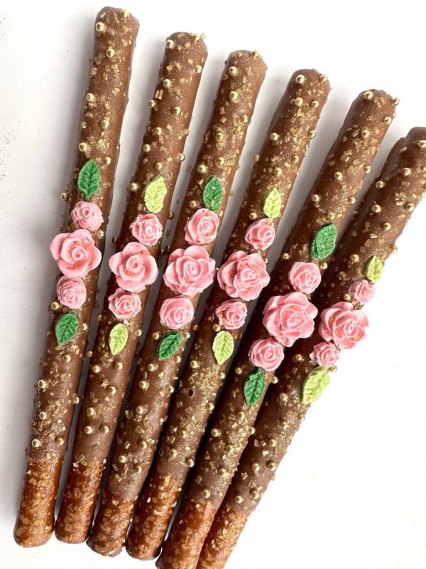 Five milk chocolate-covered pretzel sticks, adorned with pink rose and green leaf icing, placed side by side on a white background; part of the 12-Dainty Pretzels collection for birthdays, weddings, or shower favors.