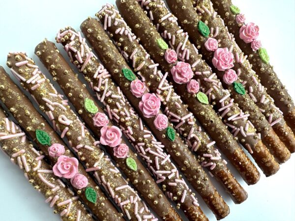 A row of decorated milk chocolate-covered pretzel sticks features pink rose-shaped candies, green leaves, and pink and gold sprinkles on a white surface. Perfect for birthday parties, weddings, or shower favors.