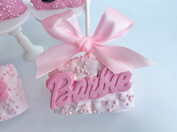 A 12-pack of pink, Barbie-themed chocolate-covered Rice Krispie treats features a square dessert with white icing, adorned with small pearls and topped with a large pink "Barbie" sign and a pink bow.