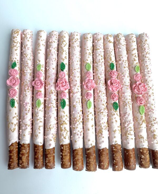 Twelve pale pink chocolate-covered pretzel rods, ideal for birthday parties, weddings, or showers, are adorned with floral and gold designs and arranged in a row on a white surface.