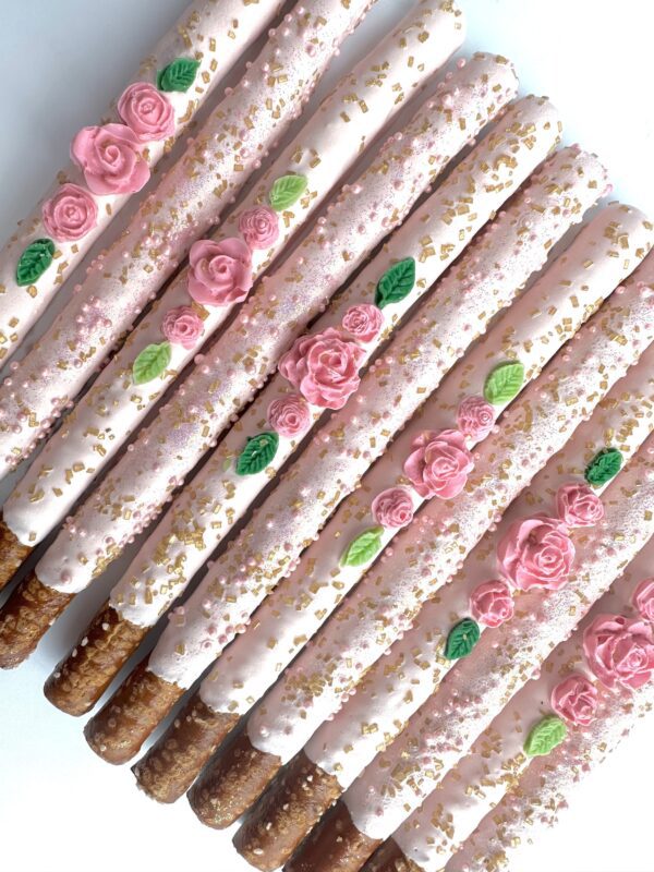 12 pale pink pretzel rods coated in chocolate, adorned with pink and green floral decorations and gold sprinkles, arranged on a white background; perfect for parties or favors.