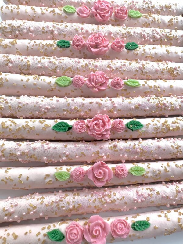 An arrangement of pretzels covered in white icing with pink rose designs, adorned with green leaves and gold flakes, ideal for birthday parties or wedding and shower favors.