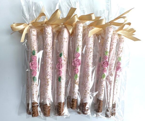 A row of the 12-Pale Pink Chocolate Covered Pretzels, adorned with pink roses and gold sprinkles, each individually wrapped in clear plastic and tied with a gold ribbon, perfect for birthday parties, weddings, or shower favors.