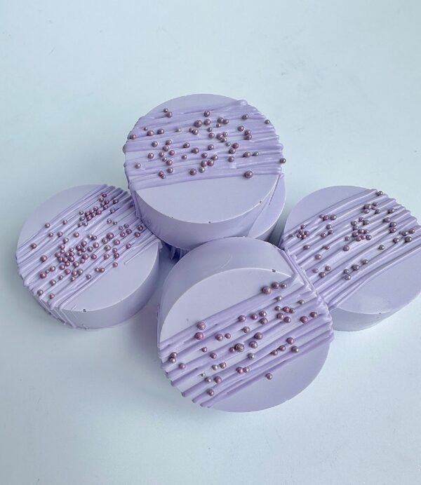 Four lavender-colored, drizzled chocolate-covered Oreos with bead details are arranged on a light surface.