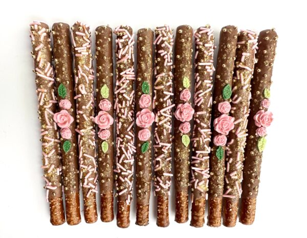 Twelve milk chocolate-covered pretzel rods featuring pink roses, sprinkles, and leaves arranged on a white backdrop. Ideal for birthdays, weddings, or showers.