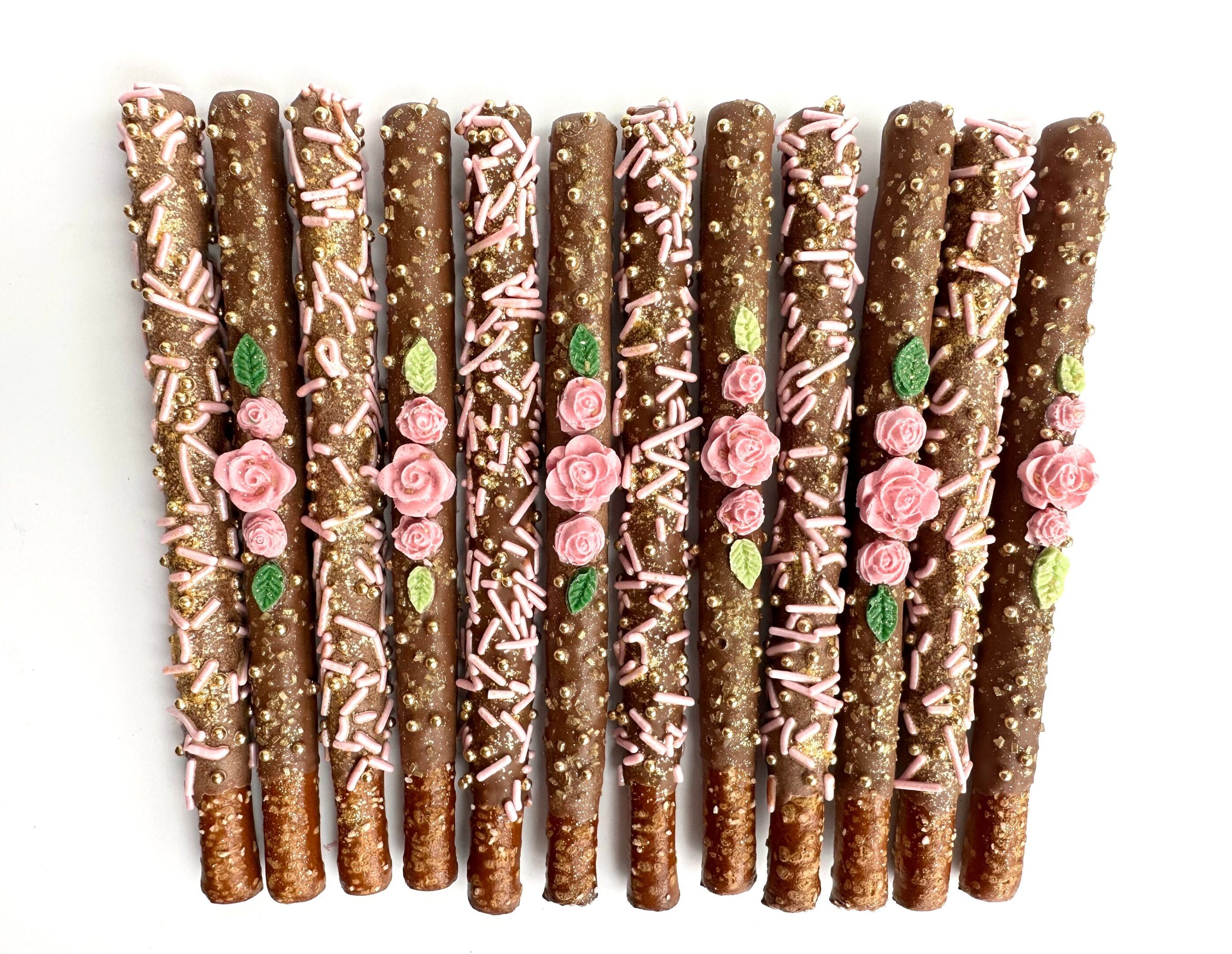 Twelve milk chocolate-covered pretzel rods featuring pink roses, sprinkles, and leaves arranged on a white backdrop. Ideal for birthdays, weddings, or showers.