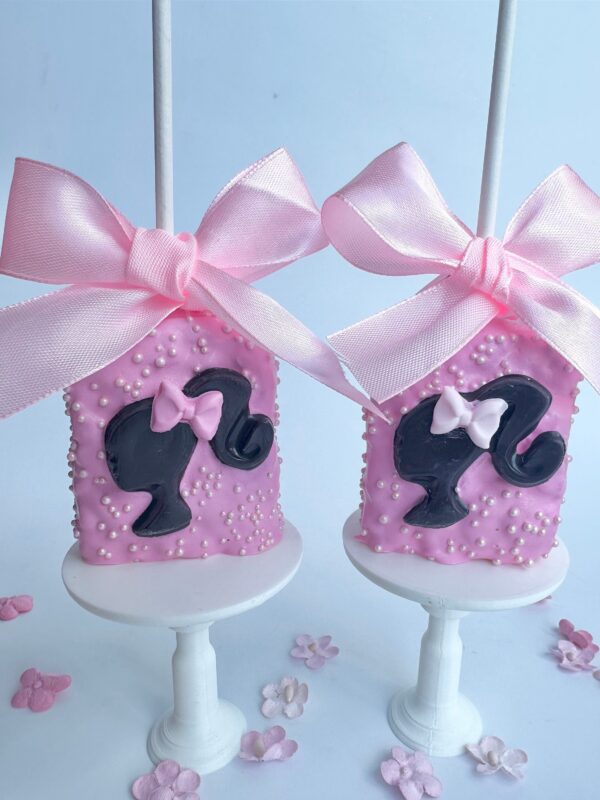 Two pink Barbi-themed chocolate-covered Rice Krispie treats feature a girl's head in black icing, topped with large pink bows and scattered with small white pearls.