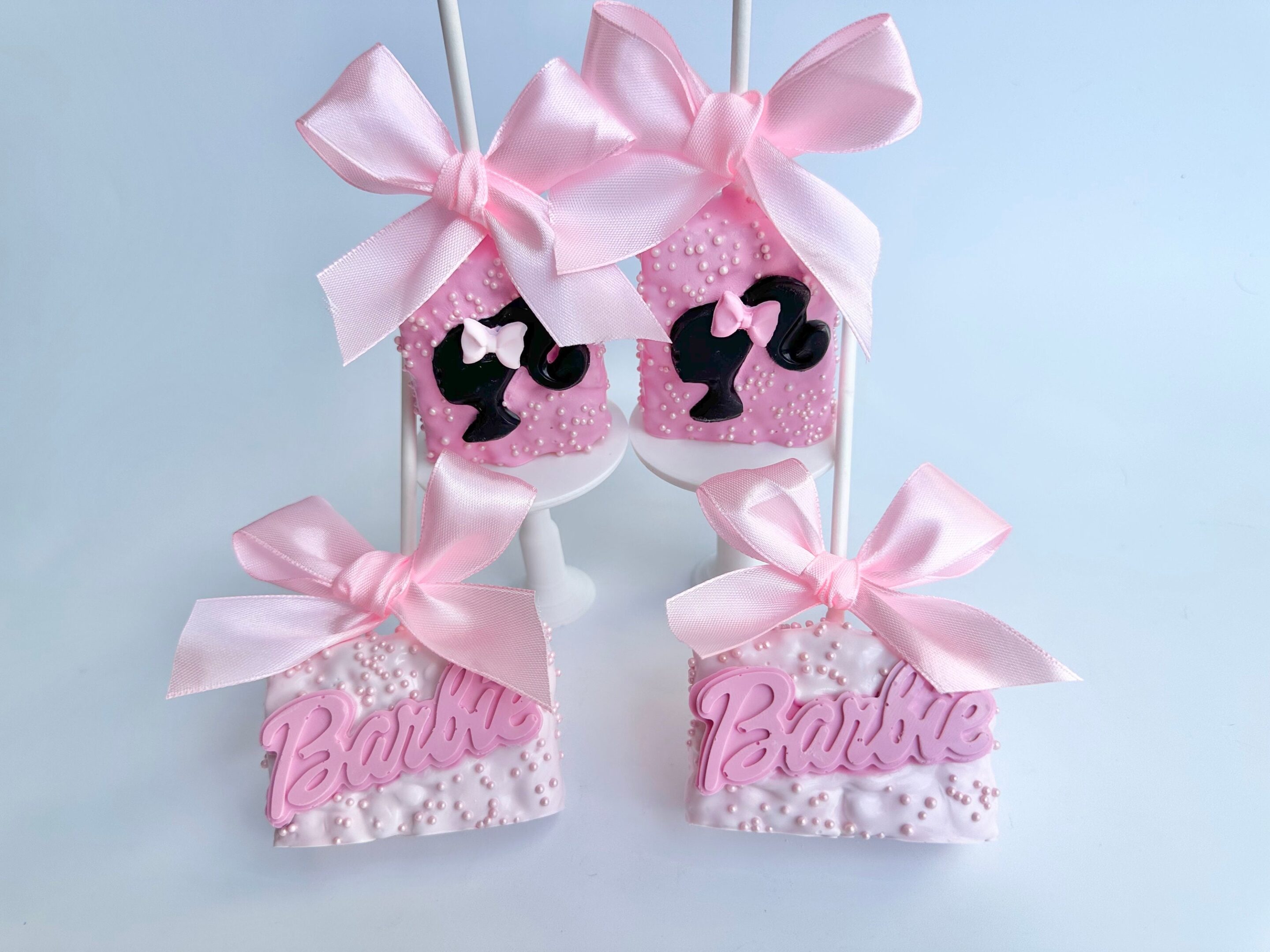 12 pink Barbi-themed Rice Krispie treats, decorated with "Barbie" and silhouette designs plus large pink bows on top, are displayed on a stand.