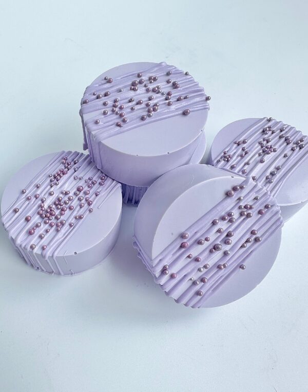 Four lavender-colored chocolate-covered Oreos, adorned with sprinkles and drizzle, are placed on a light surface.
