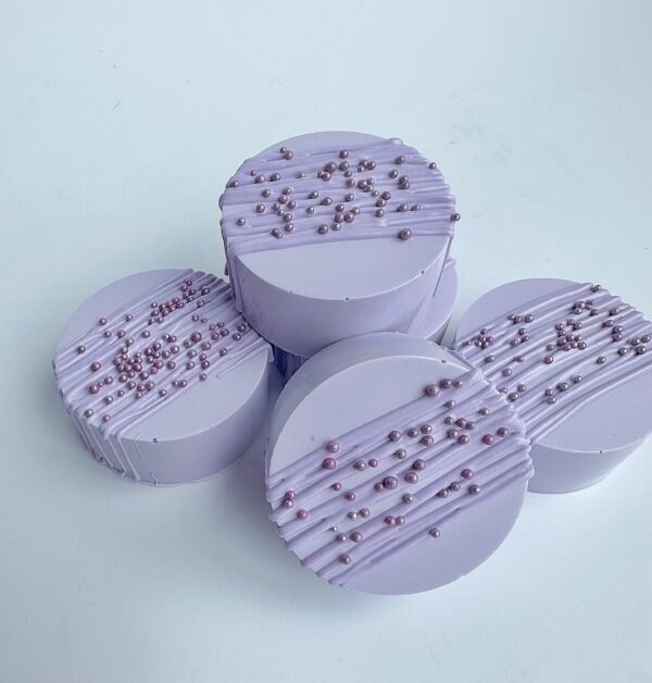 Four lavender-colored chocolate-covered Oreos with diagonal lines and small pearl decorations are displayed on a white background.