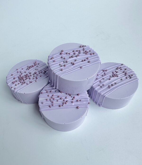 A set of four round lavender chocolate-covered Oreos with drizzled icing and metallic pearl decorations, displayed on a white background.